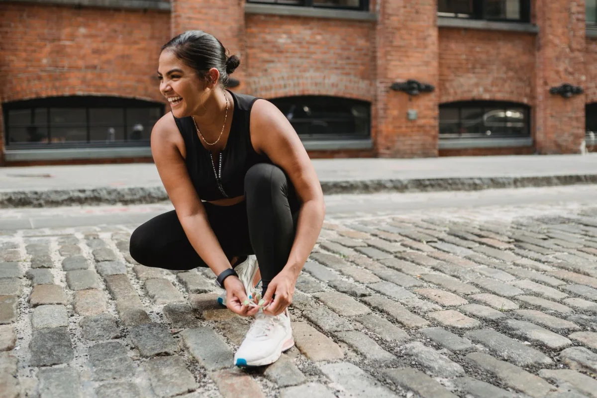 Runner crouching to tie her shoes on a cobbled path beside a brick building.